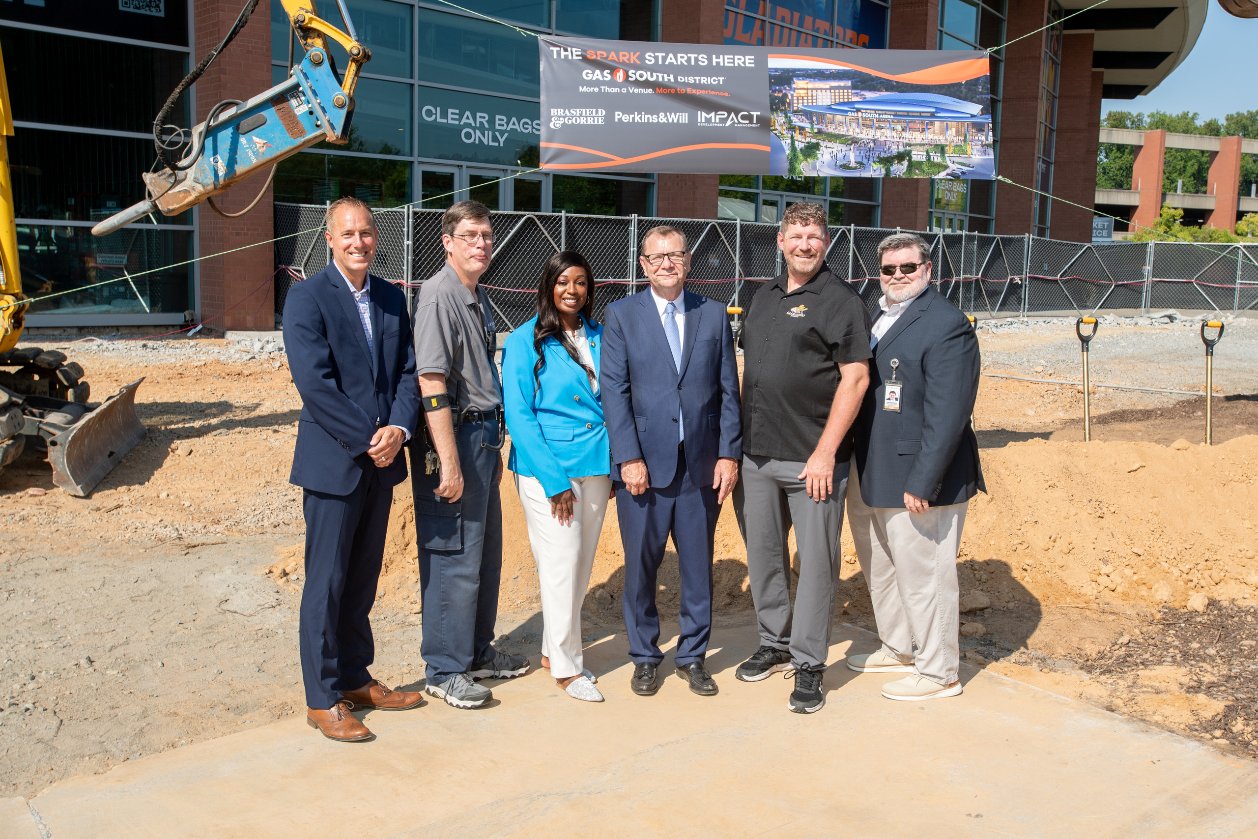 Six people stand at the Gas South Arena renovation groundbreaking site, with construction equipment and a rendering of the redesigned arena behind them.